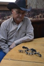 Larry Bacon sits at a table with a pair of silver spurs and wears a blue shirt and blue cowboy hat.