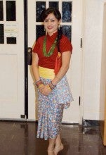 Nitu Rajbahndari stands in front of a white door and wears a red shirt, blue floral skirt, yellow fabric belt, and a green necklace.