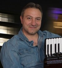 Milen Slavov sits with a black accordion in front of a dark blurred background. He wears a long sleeved denim shirt.