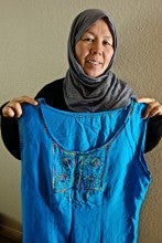 Basira Sadiqi stands holding a blue embroidered dress. She wearing a gray hijab and a black sweater.