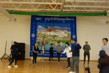 Members of the Northwest Tibetan Cultural Association dance together in a room with white walls and a large blue tapestry that says "Northwest Tibetan Cultural".