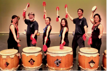 Monmouth Taiko stand on a stage with a beige background and pose with red drumsticks behind five taiko drums. All members are wearing black shirts and black pants.