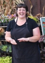 Sandra Porter stands outside in front of shelves with potted plants. She is wearing a black shirt with a black apron.