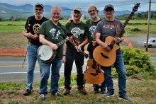 The Brownsmead Flats pose outdoors with two acoustic guitars, a banjo, and a fiddle with greenery and hills in the background. They are wearing t-shirts and jeans.