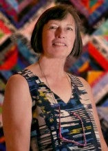 Theresa Richards Chickering stands in front of a colorful geometric quilt on the wall. She is wearing a patterned tank top.