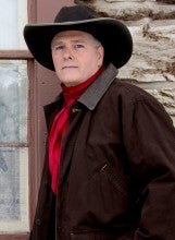 Tom Swearingen stands next to a wooden building wearing a black jacket, a black cowboy hat, and a red scarf.