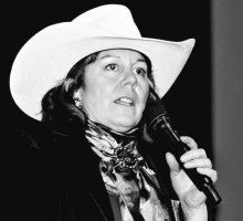 A black and white image of Kathy Moss wearing a cowboy hat and speaking into a microphone.