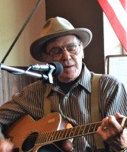 Ron Phillips sits playing an acoustic guitar and singing into a microphone. He is wearing a tan hat and a blue and white striped button down shirt with suspenders.