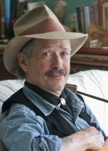 Mark Ross sits on a white couch and holds an acoustic guitar. He wears a blue collared shirt and tan hat.