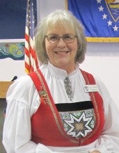 Patti Jo Meshnik stands next to a table displaying her rosemåling work. She wears a traditional Norwegian dress with a decorative collar and a red vest.