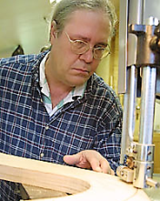 Tommy Nunn uses an electric saw machine and works on a wooden instrument. He wears a blue plaid shirt.