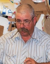 Steve McKay sits at a workbench with a leather saddle piece held down with his left hand and a hammer in his right. He is wearing a white striped button-up shirt.