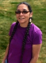 Sara Barton stands next to a table with an article of Native American regalia made from leather and decorated with small beadwork. She is wearing a purple shirt, blue jeans, and sunglasses.