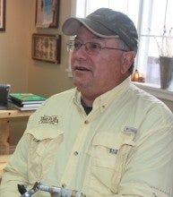 John Krueger sits indoors in front of a metal tool. He wears a pale yellow collared shirt and a gray baseball cap.