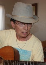 Lisa Ackerman plays a brown guitar while sitting inside and wearing a pale yellow shirt, gray hat, and jeans.