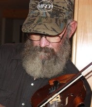 Larry McLain stands indoors and plays a fiddle. He wears a black collared shirt and a camo baseball cap.