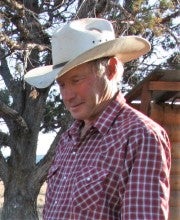Keith Barnhart stands outside with trees and a small wooden building in the background. He wears a red plaid buttoned shirt and a white cowboy hat.