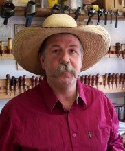 Len 'Peanut' Babb stands in front of racks of leatherworking tools. He wears a wide brimmed straw hat and a red collared shirt.