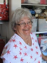 Lois Kropf in a white shirt with stars sitting in front of shelf of quilting supplies.