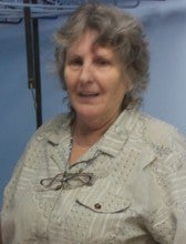 Linda Wood stands in a room with blue walls. She wears a gray/beige collared shirt.