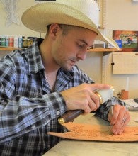 Joe Meling sits working on an adorned piece of leather. He wears a blue plaid collared shirt and a white cowboy hat.