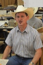 Richard Stapleman sits in his workshop. He wears a blue and white striped button-down shirt and a white cowboy hat.