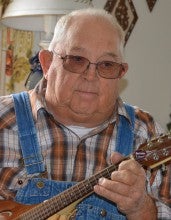 Robert (Bob) Stevens sits in an armchair holding a mandolin. He wears a brown plaid shirt and blue overalls.