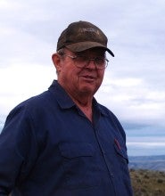 Sidney "Sam" Seale stands outdoors with a cloudy sky and hills in the backgrund. He is wearing a blue button-up shirt, blue jeans, and a brown baseball cap. 