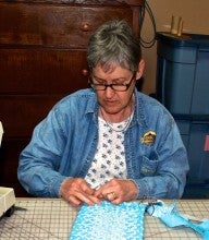 Judi Galvin sits at a table next to a sewing machine and weaves a small blue rug. She wears a denim shirt over a white floral shirt. 