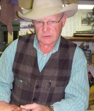 Ron Wilson sits at a table near a workbench with a piece of leather. He wears a beige cowboy hat and a grey plaid vest over a blue shirt.