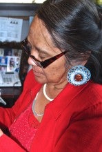 Marjorie Kalama sits at a desk and works on beadwork. She wears a red and white polka dot shirt, a red blazer, and large round beaded earrings. 