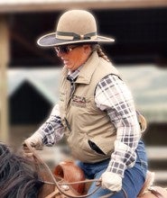 Kitty Lauman rides a brown horse in an outdoor arena. She wears a beige brimmed hat, plaid shirt, beige vest, and blue jeans.