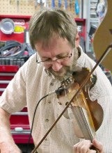 Steve Campbell stands and plays an electric fiddle in his workshop. He is wearing a white button-up shirt and blue jeans.