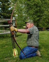 James Akenson kneels while shooting a bow and arrow and wears a gray shirt, camouflage hat, and blue jeans.