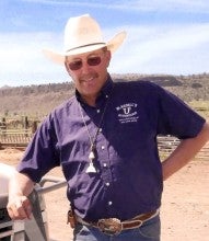 Tom Blasdell stands outdoors next to a white truck. He is wearing a purple shirt that says "Blasdell's Stockdogs", blue jeans, and a white cowboy hat.