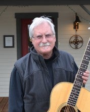 Preston Thompson stands outside his guitar shop holding an acoustic guitar. He wears a black vest over a black long sleeved shirt.
