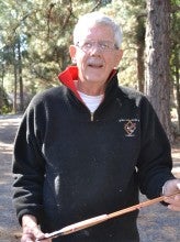Roger Fairfield stands outdoors and holds a bamboo fly fishing rod with tall trees in the background. He wears a black jacket and jeans.