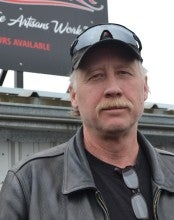 Steve Parks stands outside in front of the Parks Bronze sign. He is wearing a black shirt, a black leather jacket, and a black baseball cap.