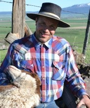Pat Dougherty sits outside against a rock jack style fence with a dog on his lap. He wears a checkered button up shirt and a black brimmed hat.