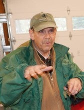 Ron Lauzon stands inside a garage next to a river raft boat. He wears a green jacket and a green baseball cap.
