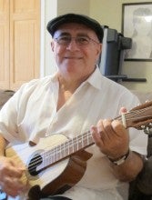 Neftali Rivera sits on a gray couch wearing a white shirt, black hat, and jeans. He plays a small acoustic guitar.
