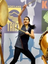 Kelsey Furuta stands on a stage and energetically plays two large taiko drums. She wears a blue wrap shirt and dark pants.