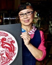 Judy Zhou holds a red and white papercut art piece. She wears a pink long sleeve shirt under a blue corduroy apron.