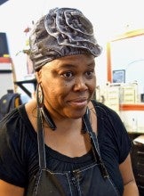 Secret Bryant stands in her Portland studio, "Southern Styles & Barber," braiding pink extensions. She is wearing a black shirt, a black apron, and a black bonnet with a flower on it.