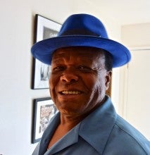 Norman Sylvester stands in front of a white wall with two hanging picture frames. He wears a blue collared shirt and blue fedora.