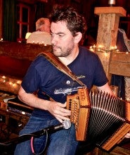 Johnny B. Connolly sits and plays a button box accordion in front of a microphone. He wears a dark blue t-shirt and blue jeans.