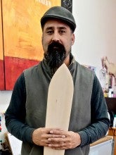 Shirod Younker stands next to a large yellow and red painting with a wooden canoe oar. He is wearing a gray vest, a black long-sleeved shirt, dark blue jeans, and a black hat.