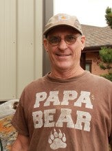 Kevin Strauslin stands outside of his shop and wears a brown t-shirt that says "papa bear" and a light brown baseball cap.