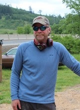 James stands outside leaning against a wood chunk and wears a long sleeve blue shirt, gray baseball cap, and black pants.