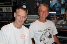Two men sit in a music studio in front of large mixers and both wear white t-shirts.